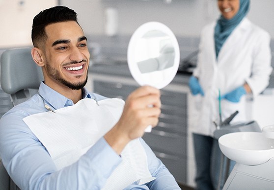 Man in dental chair smiling into handheld mirror with dentist blurry in background